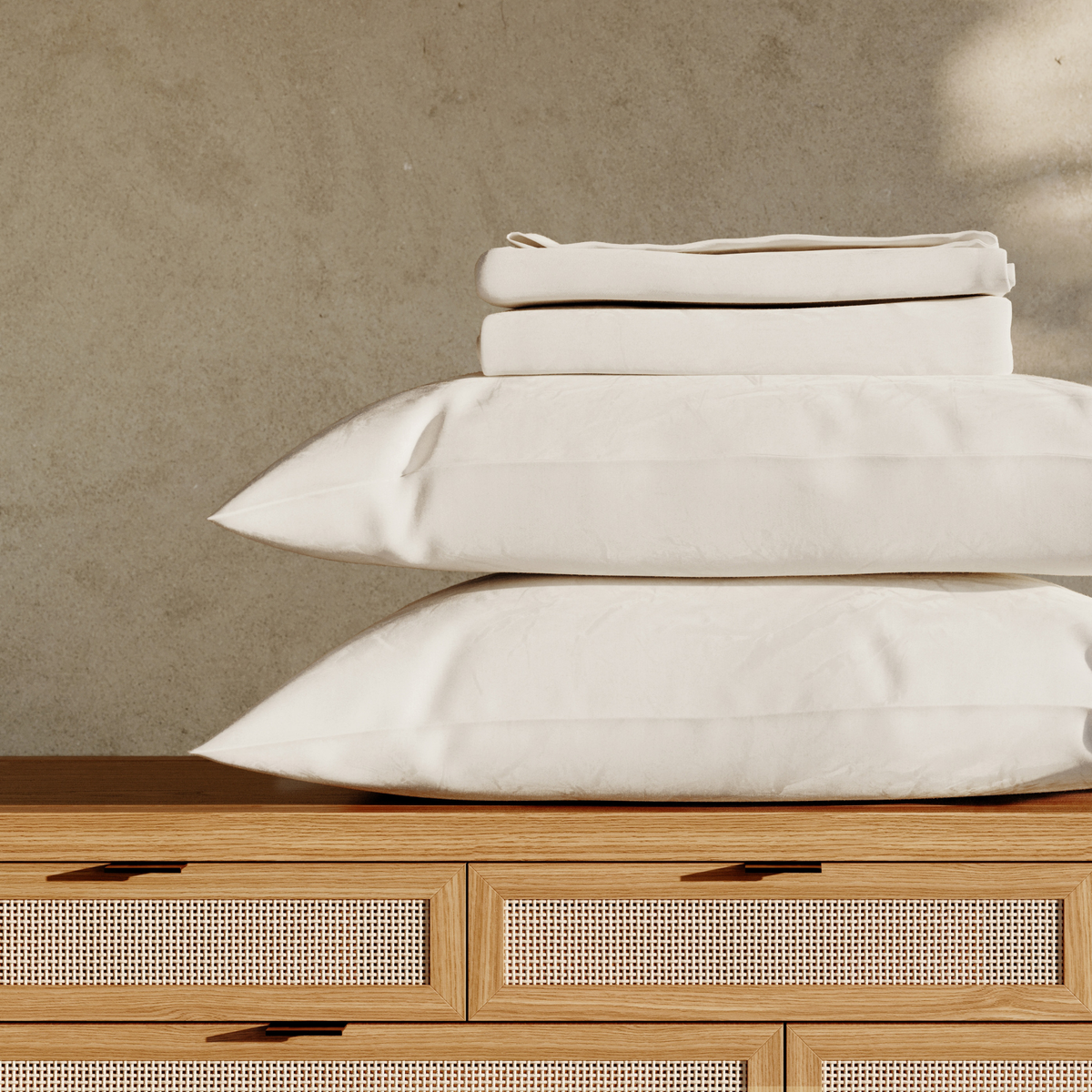 Stack of white pillows and folded sheets on a wooden dresser with a neutral background #color_arctic snow