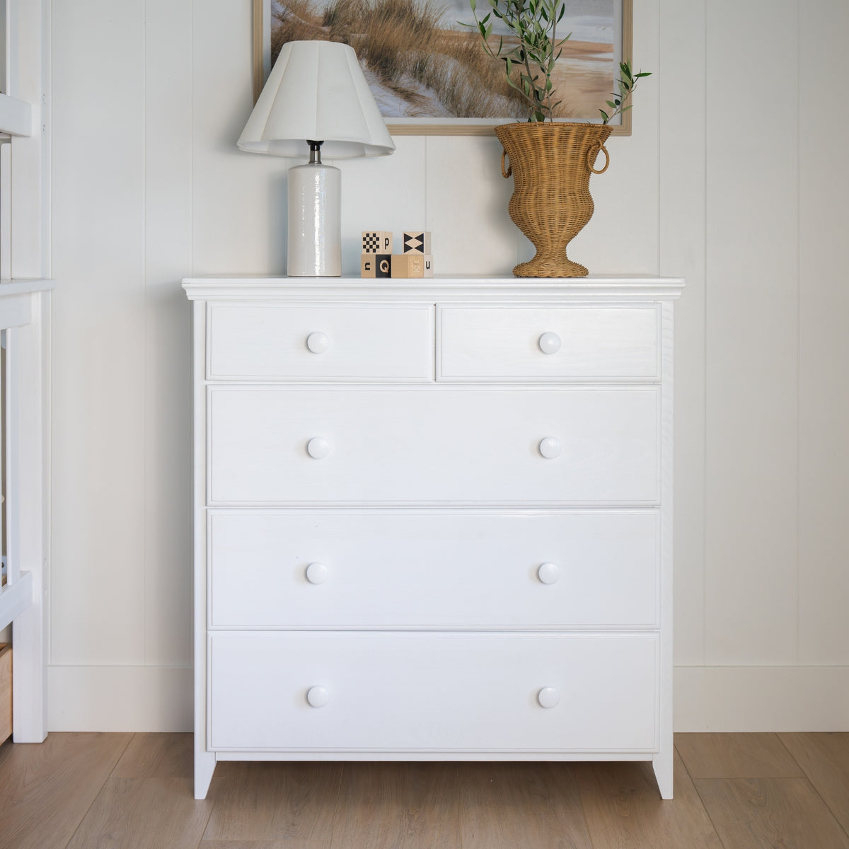 A white wooden five-drawer dresser with round white knobs, styled with a lamp, small decorative blocks, and a wicker vase  #color_washed white