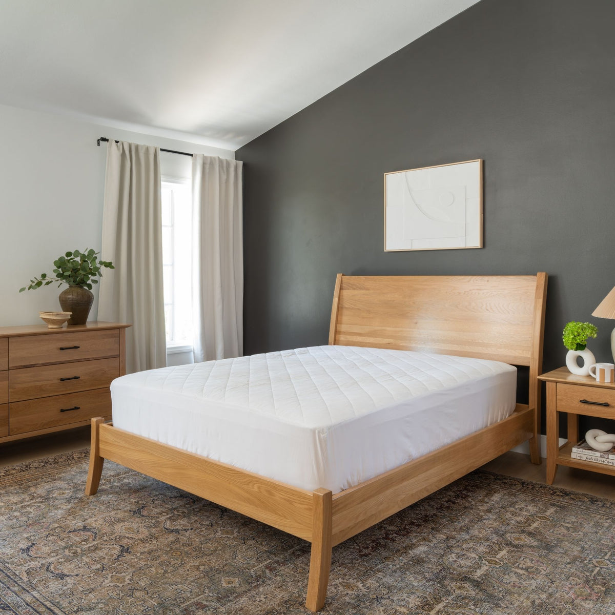 A light oak wood bed frame with a white quilted mattress pad, styled in a modern bedroom with a dark gray accent wall, beige curtains, and a patterned rug.